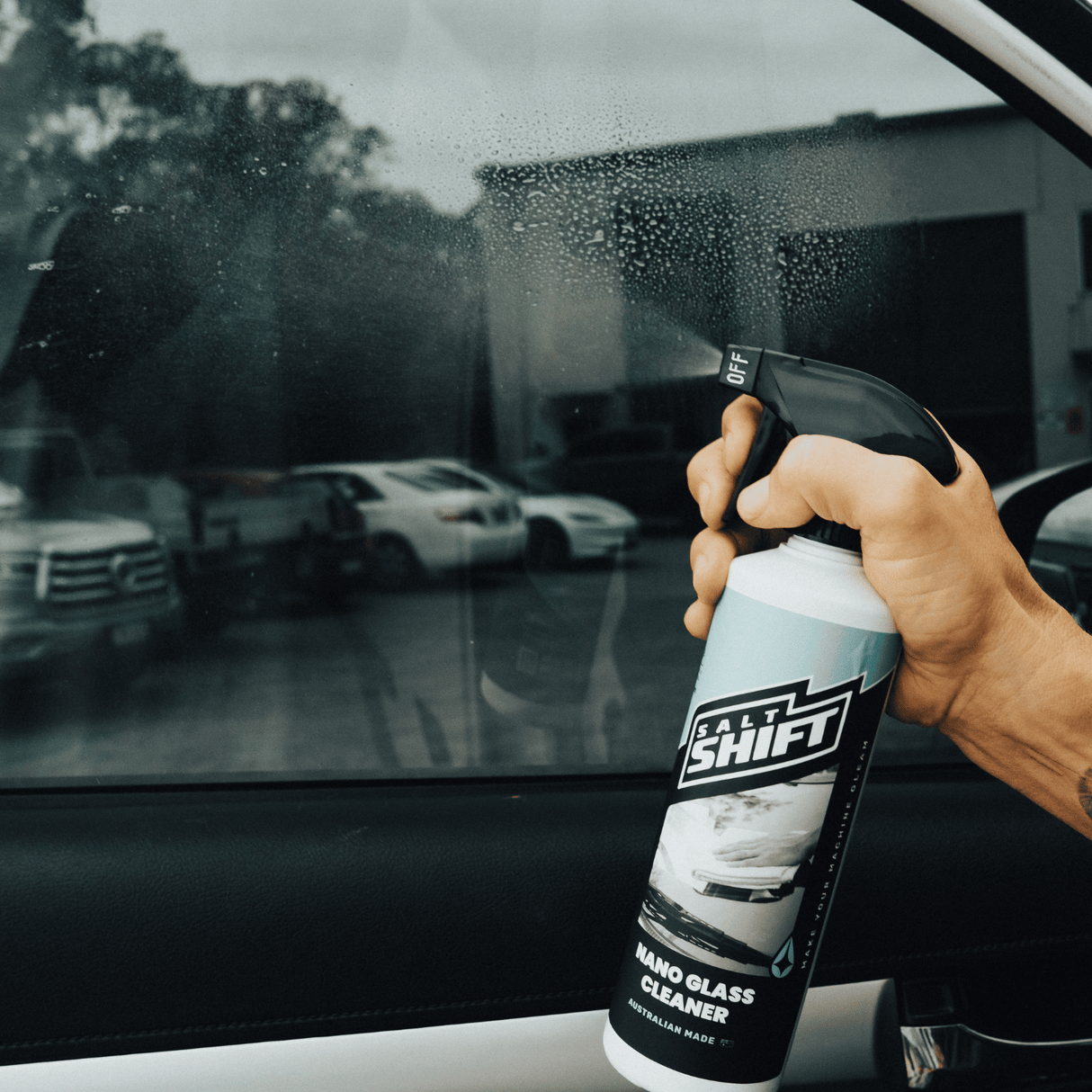 Hand spraying Salt Shift Nano Glass Cleaner on a car window to remove dirt and streaks, ensuring a crystal-clear finish. 